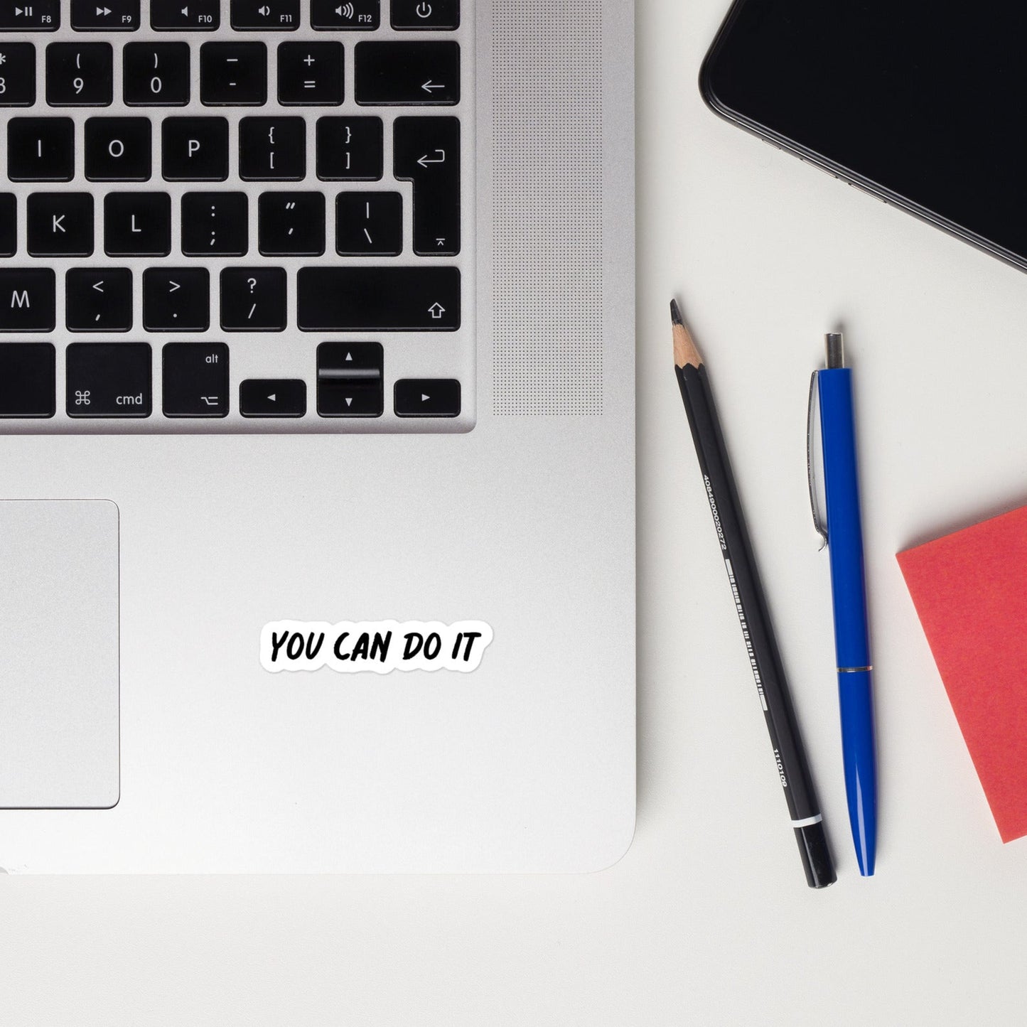 A laptop with a motivational sticker that says 'YOU CAN DO IT' positioned on the lid, alongside a mobile phone and a pencil.
