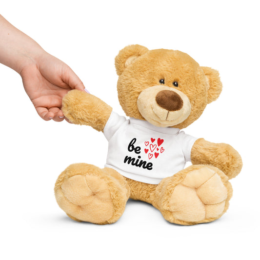 A stuffed teddy bear with a white t-shirt that says 'BE MINE' and has heart illustrations holding a hand.