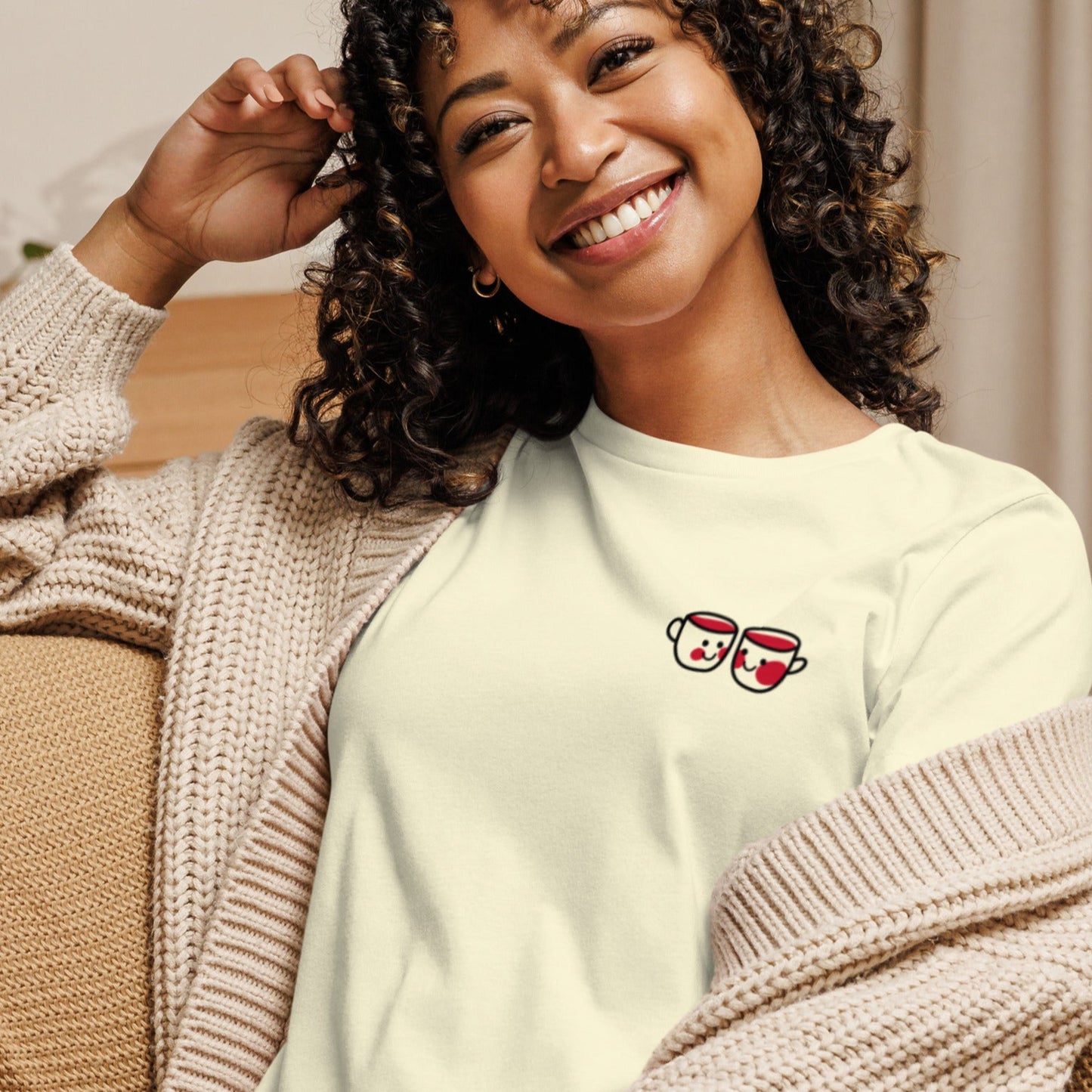 A smiling woman with curly hair wearing a white crew neck t-shirt with with two hot chocolate mugs, paired with a beige cardigan, sitting on a couch.
