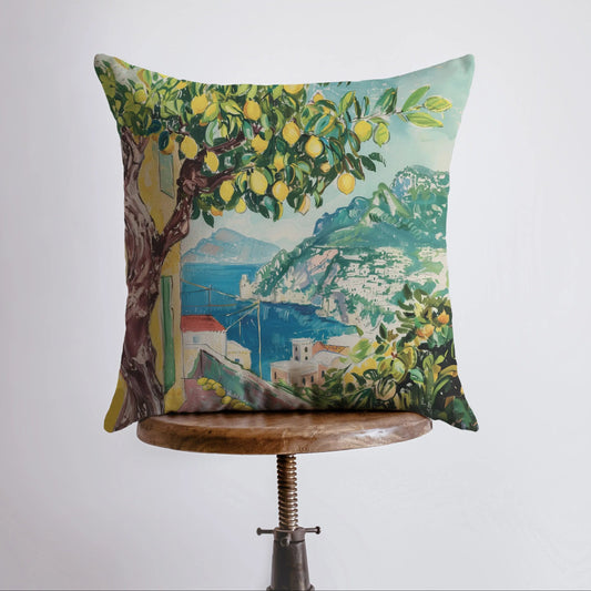 Decorative pillow with a scenic design of a lemon tree and village on a wooden stool against a white background.