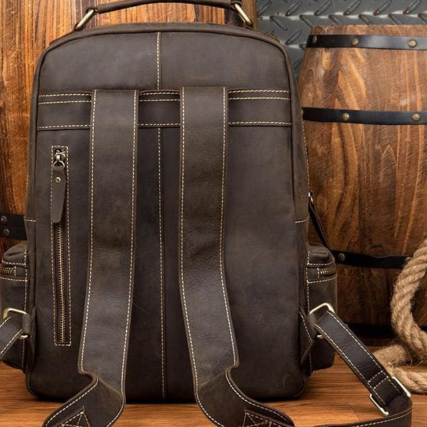 Brown leather backpack on a wooden surface with barrels and a bottle in the background