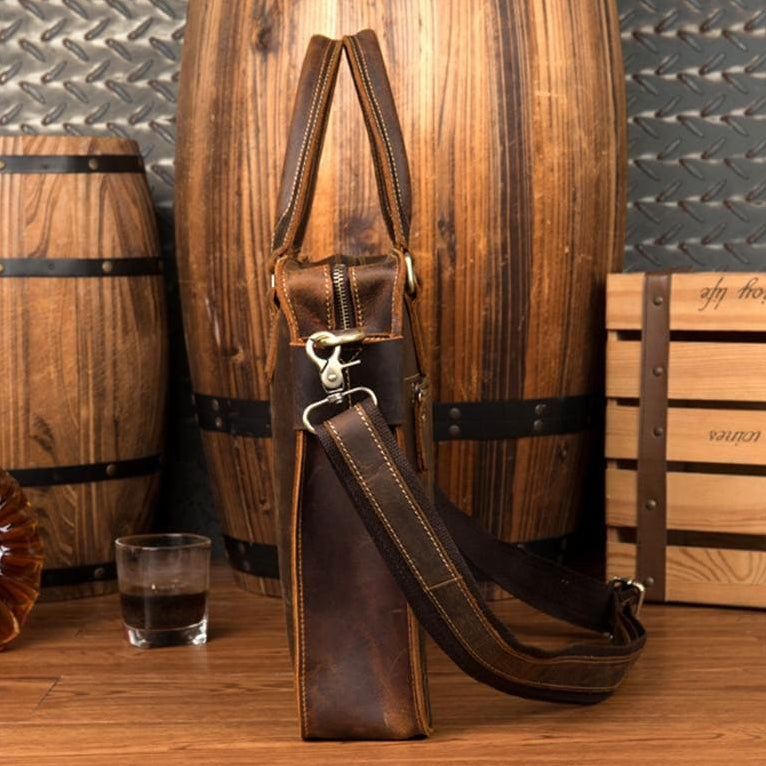Brown leather bag with a glass of whiskey on a wooden surface with barrels in the background